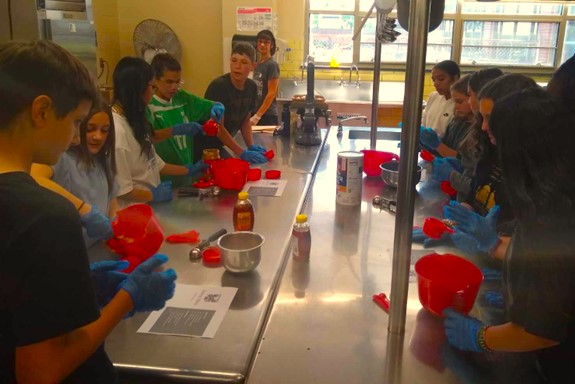 students holding measuring cups along a stainless steel counter in a school kitchen