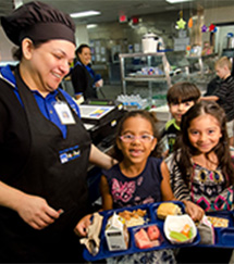 a school meal professional standing next to two smiling girls holding lunch trays