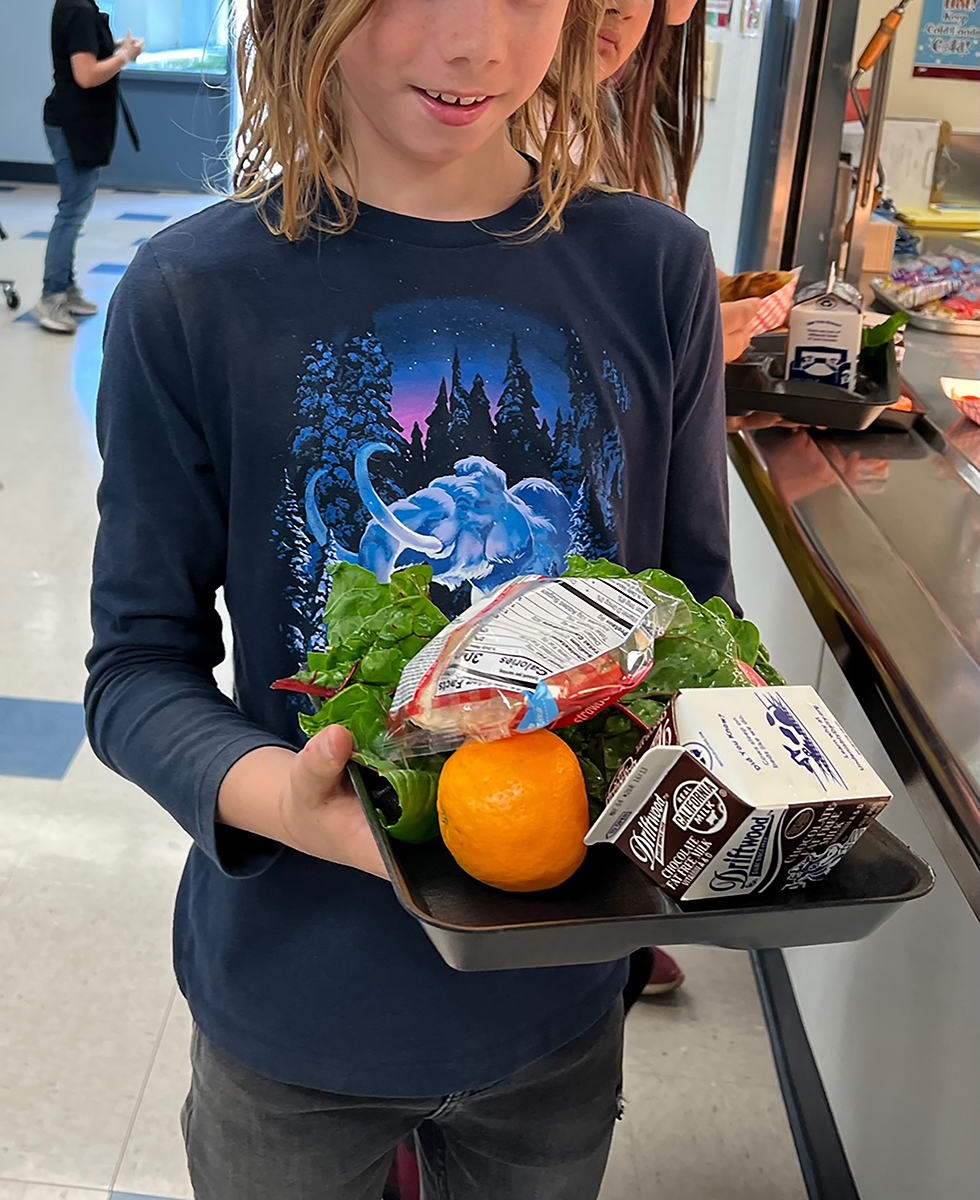 A child holding a tray of food with rainbow chard, an orange, and chocolate milk.