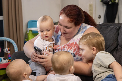 a woman looking at her phone holding an infant and surrounded by 3 toddlers
