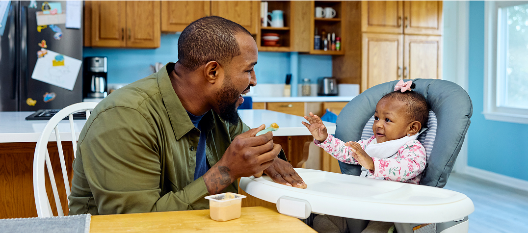 man in a kitchen feeding a baby girl in a highchair