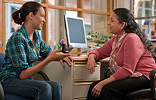 two women talking at a desk in front of a computer