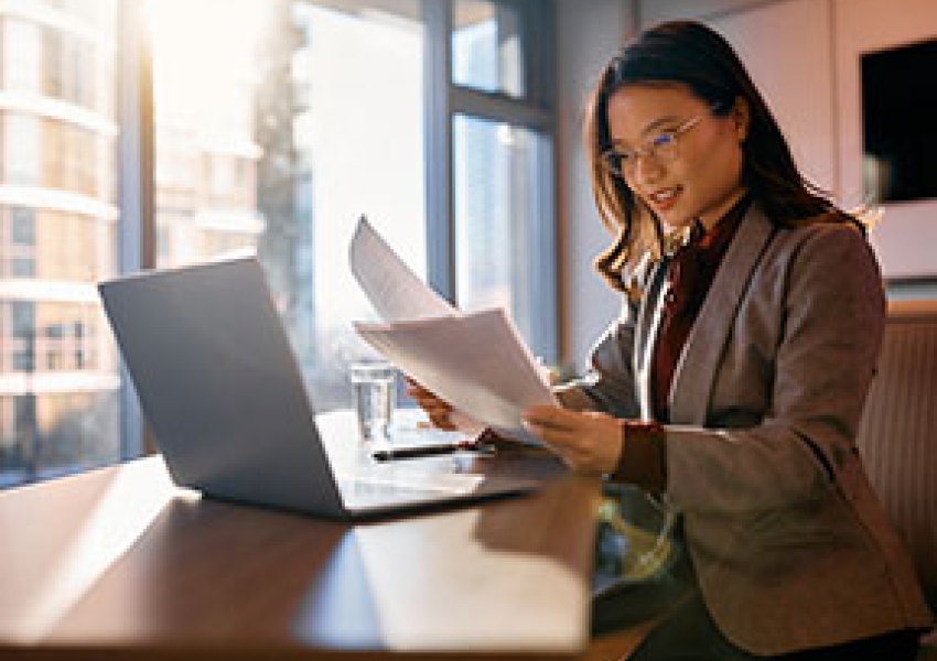 woman reading a report in front of a laptop with sun shining through a window behind her