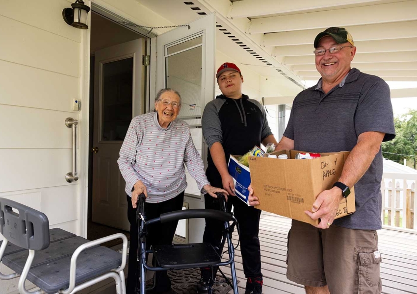 two men delivering a package of food to a senior woman using a walker on her front porch