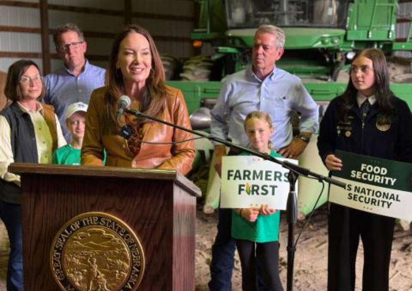 Secretary Rollins at a podium speaking with a family behind her holding signs saying Farmers First.