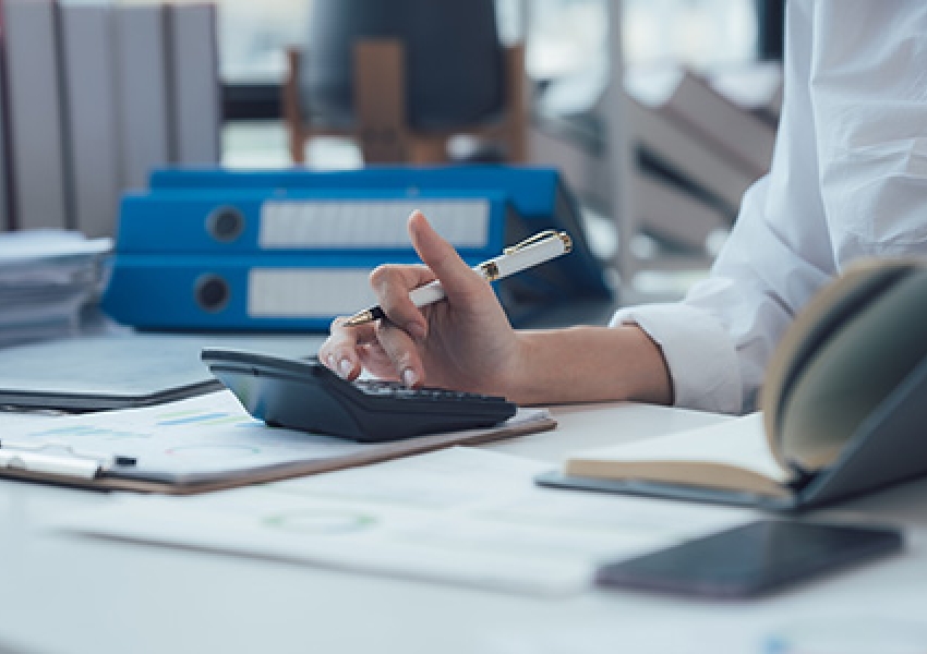 image of a hand typing into a calculator at a desk surrounded by papers and notebooks