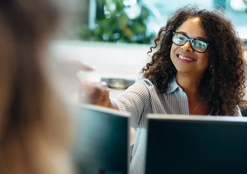woman behind a computer smiling and handing a paper over a desk