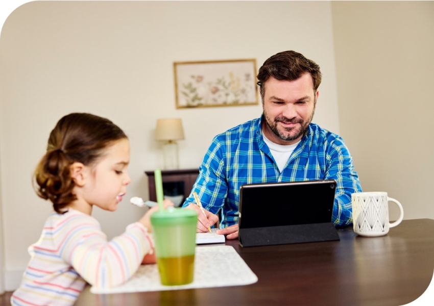 a man at a table looking at a computer with his daughter eating yogurt next to him