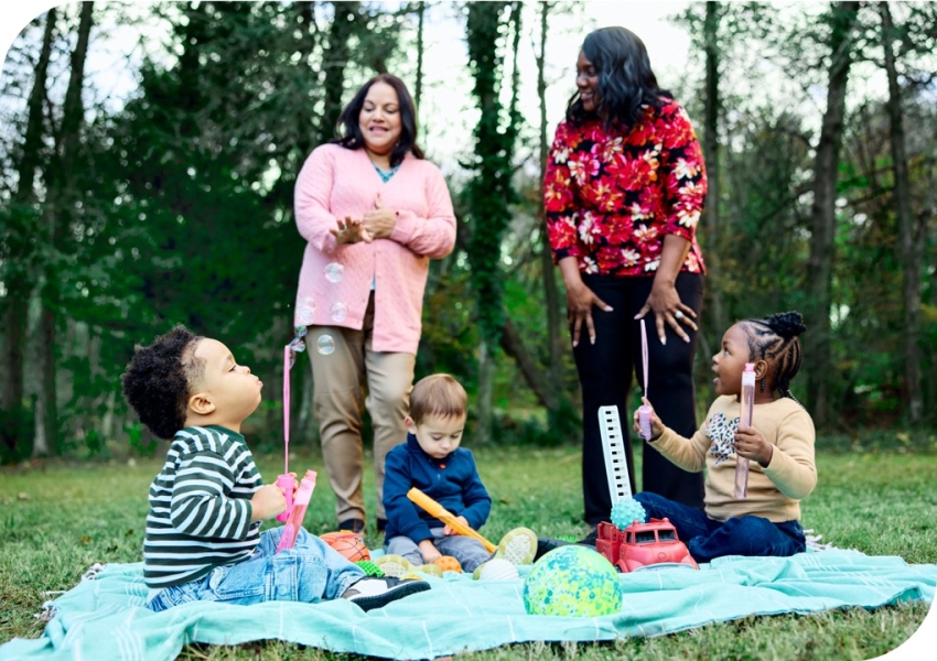 two adults standing over a picnic blanket with 3 toddlers blowing bubbles on it