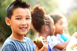 little boy eating a sandwich with other kids in background