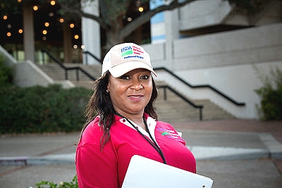 USDA FNS disaster response employee wearing a USDA ball cap, red t-shirt, and holding a clip-board in front of a building.