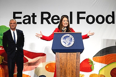 Secretary Rollins in front of an Eat Real Food sign with Secretary Kennedy behind her