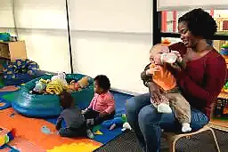 woman bottle feeding an infant while others play in the background